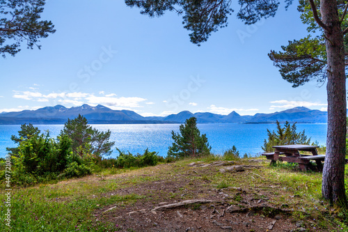 A wooden bench with benches on the bank of a picturesque wide blue river on the forest edge overlooking the mountains on the horizon under the blue sky. Norway.