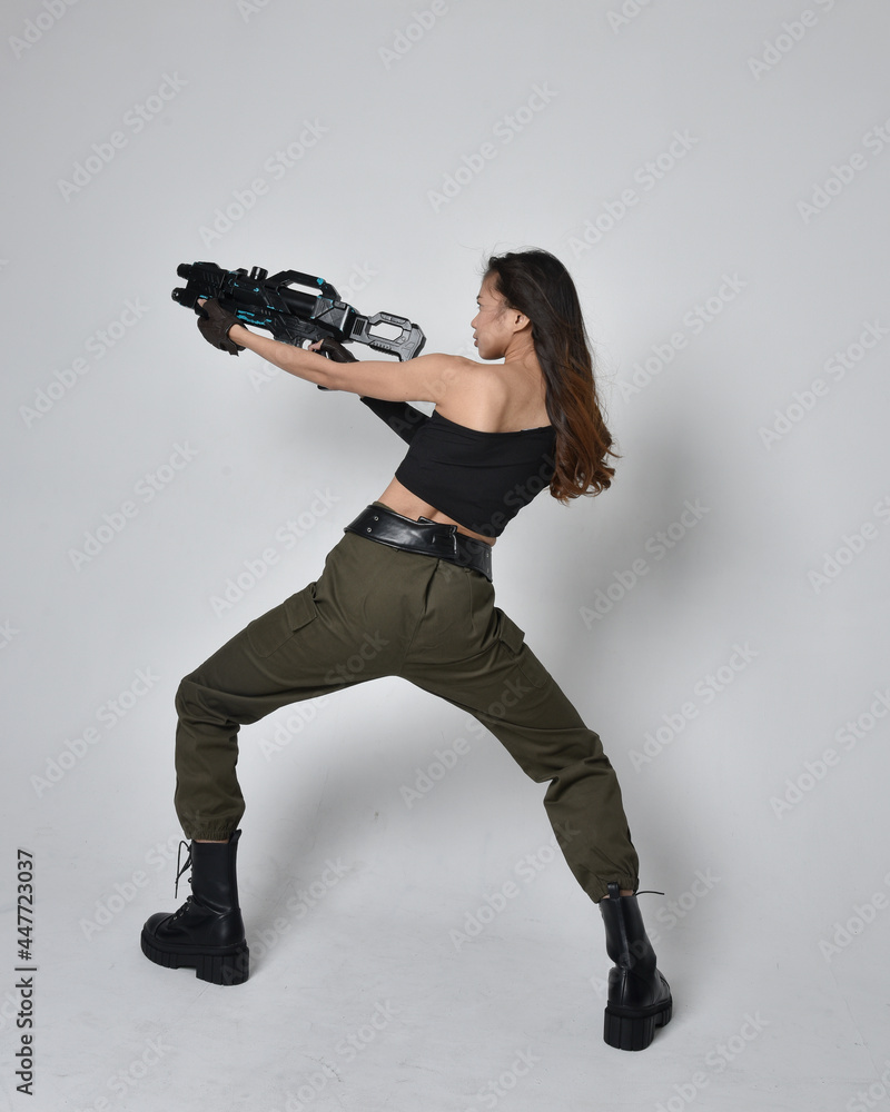 Full length portrait of pretty brunette, asian girl wearing black top and khaki utilitarian army pants and leather boots. Standing pose holding science fiction gun, isolated against studio background Full length portrait of pretty brunette, asian girl wearing black top and khaki utilitarian army pants and leather boots. Standing pose holding science fiction gun, isolated against studio background