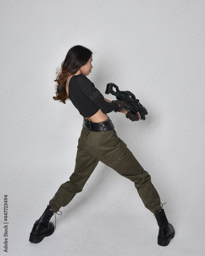 Full length portrait of pretty brunette, asian girl wearing black top and khaki utilitarian army pants and leather boots. Standing pose holding science fiction gun, isolated against studio background Full length portrait of pretty brunette, asian girl wearing black top and khaki utilitarian army pants and leather boots. Standing pose holding science fiction gun, isolated against studio background