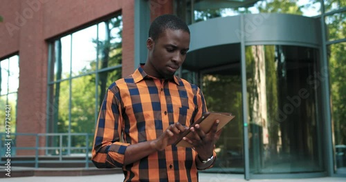 Smart dark-haired female holding tablet and searching for information for her report while standing at the street near the university. Technology and education concept