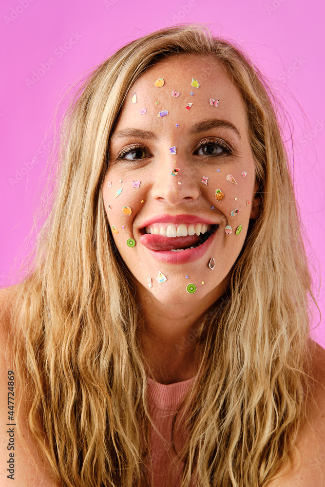 Pretty blonde girl smiling showing her tongue with colorful stickers on her face on a pink background Pretty blonde girl smiling showing her tongue with colorful stickers on her face on a pink background