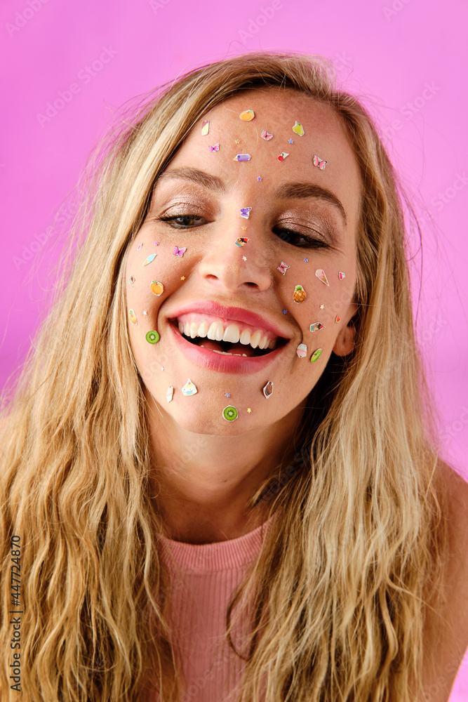 Pretty blonde girl smiling with colorful stickers on her face on a pink background Pretty blonde girl smiling with colorful stickers on her face on a pink background