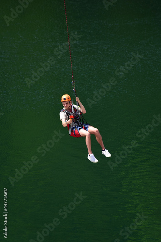 A young man in a helmet jumped from bungee jumping and is now hanging on a rope, swinging and filming himself on a sports video camera against a blurred background of water