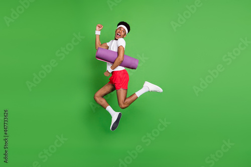 Profile photo of sporty guy jump run hold fitness mat wear t-shirt shorts sneakers isolated green color background