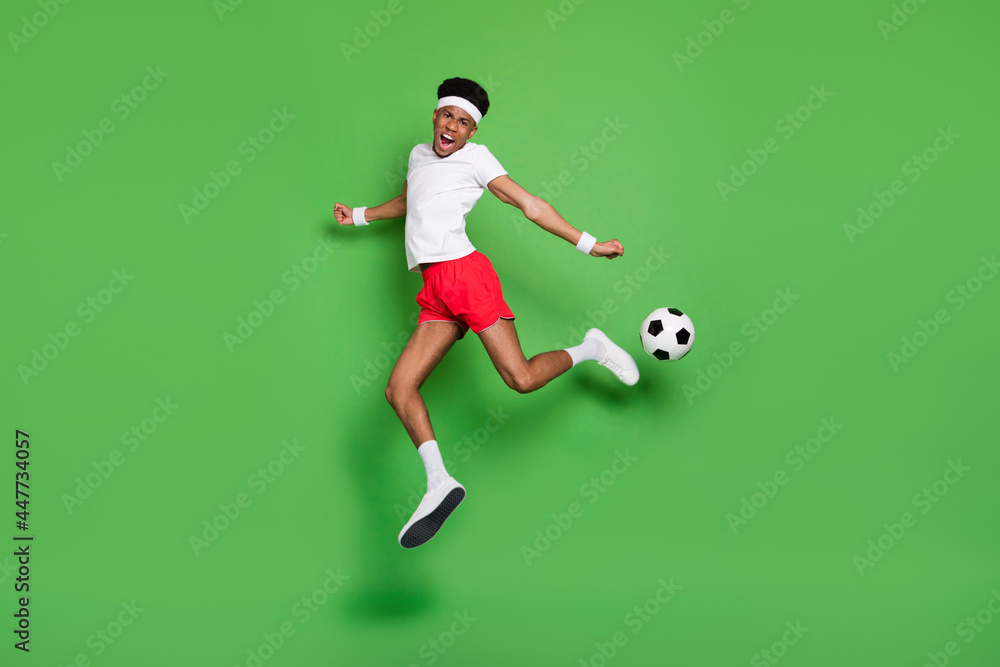 Full length photo of excited pretty dark skin guy dressed sport outfit jumping playing football isolated green color background Full length photo of excited pretty dark skin guy dressed sport outfit jumping playing football isolated green color background