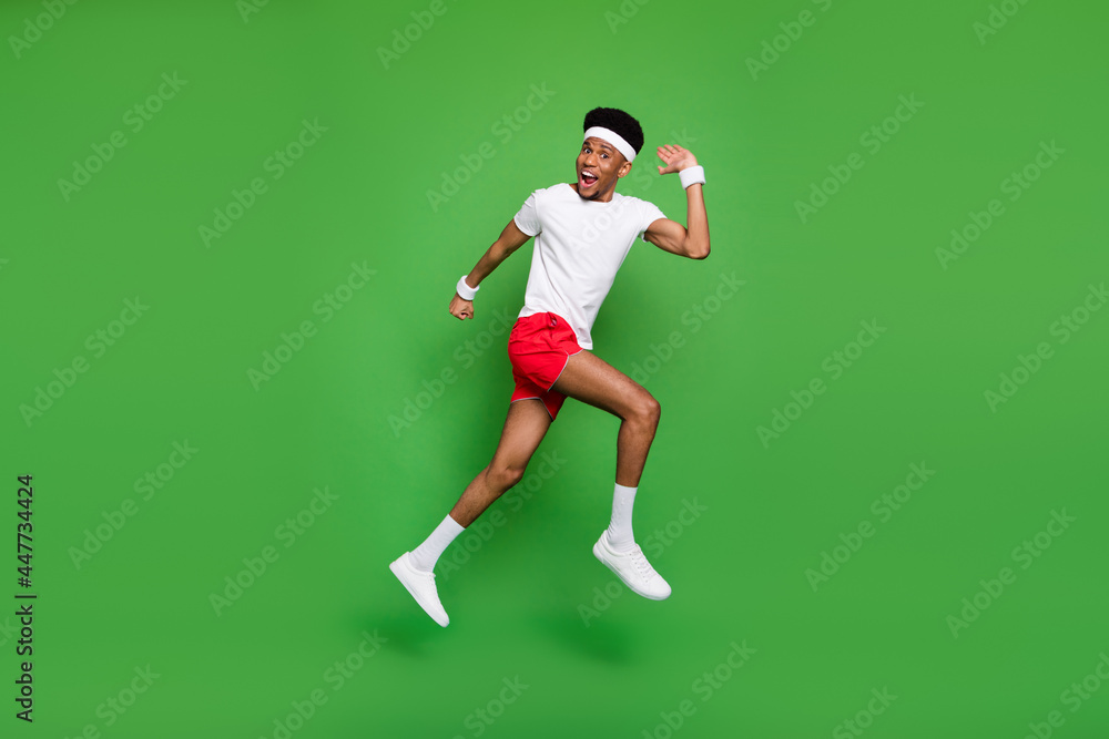 Full length photo of excited funny dark skin man wear white t-shirt jumping high running fast isolated green color background Full length photo of excited funny dark skin man wear white t-shirt jumping high running fast isolated green color background