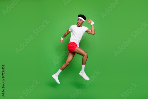 Full length photo of excited funny dark skin man wear white t-shirt jumping high running fast isolated green color background © deagreez