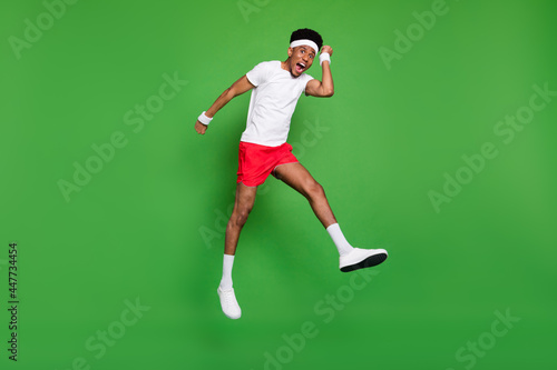 Full length photo of lucky funky dark skin guy dressed sport outfit jumping high rising fists isolated green color background © deagreez