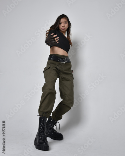 Full length portrait of pretty brunette, asian girl wearing black top and khaki utilitarian army pants and leather boots. Standing pose with gestural hands, isolated against a light grey studio backgr © faestock