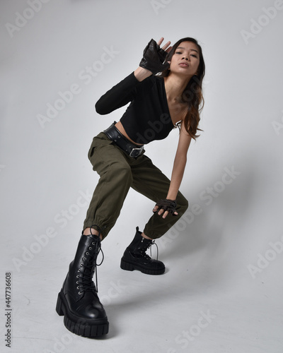 Full length portrait of pretty brunette, asian girl wearing black top and khaki utilitarian army pants and leather boots. Standing pose with gestural hands, isolated against a light grey studio backgr