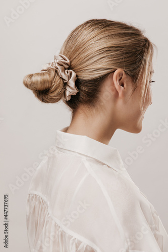 Portrait of stylish blonde with hair collected in silk beige scrunchie, wearing white blouse and posing against light background. Beauty and tenderness concept photo