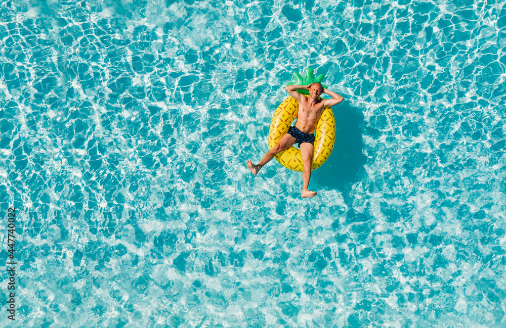 A cheerful middle-aged man in sunglasses suntanning floating on blue swimming pool waves on giant inflatable Yellow Pineapple tube and laughing. Chill out summer vacation in luxury resorts concept.