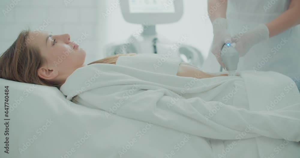 A female client is laying in a cosmetology bed in a bathrobe while a female cosmetologist is doing a SMAS lifting procedure in a stomach by using an Ultraformer apparatus. A female client is laying in a cosmetology bed in a bathrobe while a female cosmetologist is doing a SMAS lifting procedure in a stomach by using an Ultraformer apparatus.