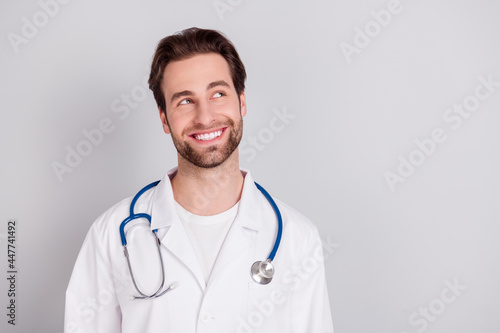 Photo of professional positive young doc man dressed white coat smiling looking empty space isolated grey color background
