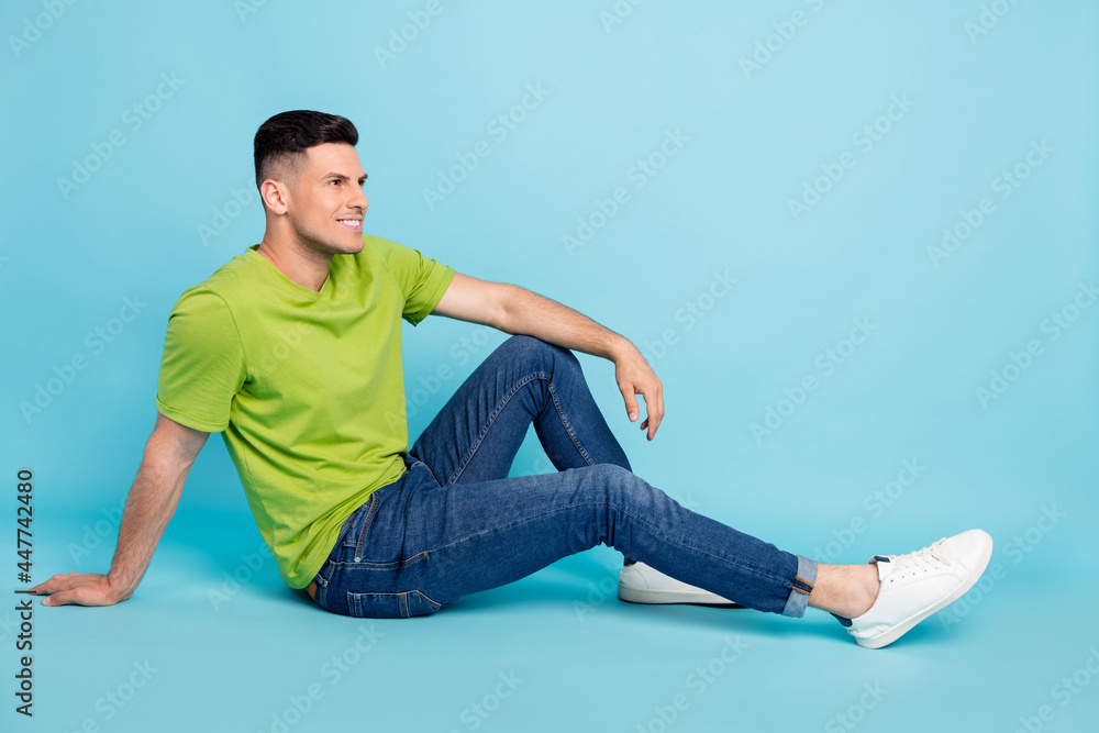 Full length photo of charming happy young man look empty space sit floor isolated on blue color background Full length photo of charming happy young man look empty space sit floor isolated on blue color background