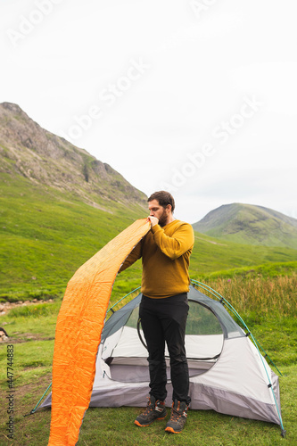 Obraz na płótnie Young and attractive man inflating a mattress next to his tent to do solo camping in Scotland. Equipment to travel during vacation w salonie