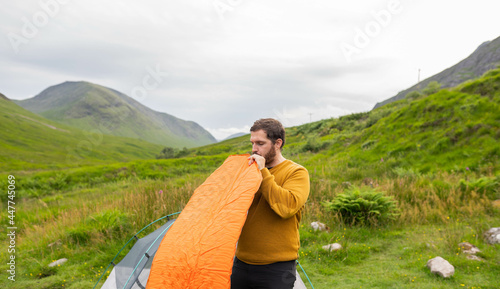 Young and attractive man inflating a mattress next to his tent to do solo camping in Scotland. Equipment to travel during vacation