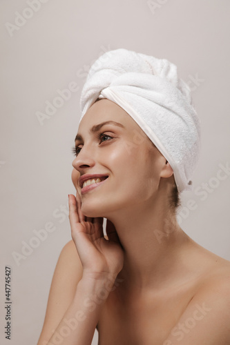 Gorgeous young lady with white towel wrapped hair and naked shoulders, smiling and looking aside isolated over light background. Body care and cleansing concept