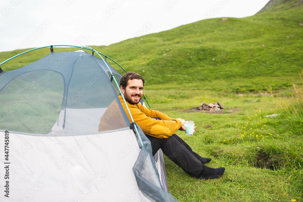 Young and attractive man sitting inside a tent outdoors enjoying a hot beverage in a reusable cup in the middle of nature in Scotland