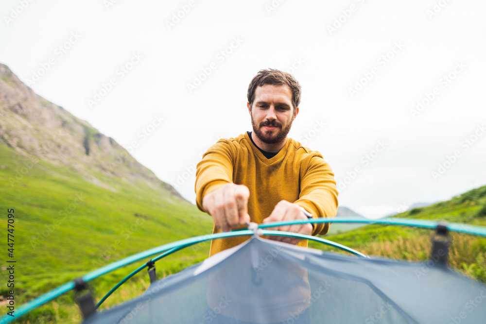 Photo of a young and attractive man setting up a camp tent outdoors to enjoy a beautiful day in the middle of nature in Scotland.