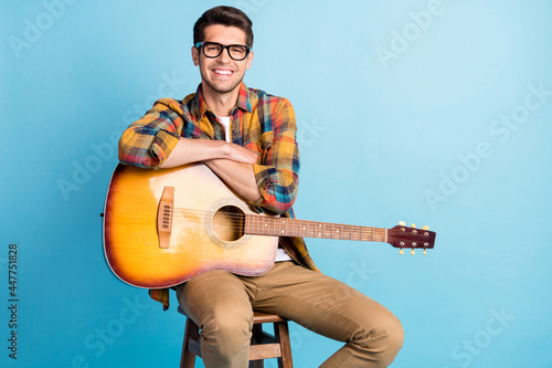 Photo of adorable cute guy dressed plaid shirt spectacles smiling siting chair holding guitar empty space isolated blue color background
