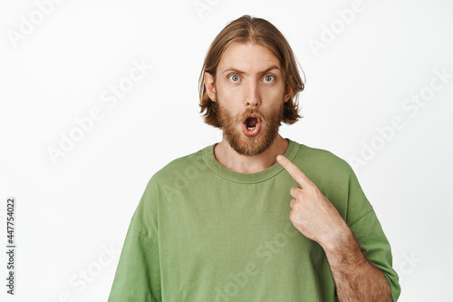 Surprised blond man stare amazed at camera, pointing finger at himself, being chosen, standing in green t-shirt against white background