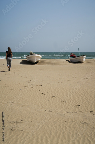 A woman silhouette seen from the back walking on a sand beach on a sunny windy day in a fishing village  two white boats on the beach and footsteps in sand