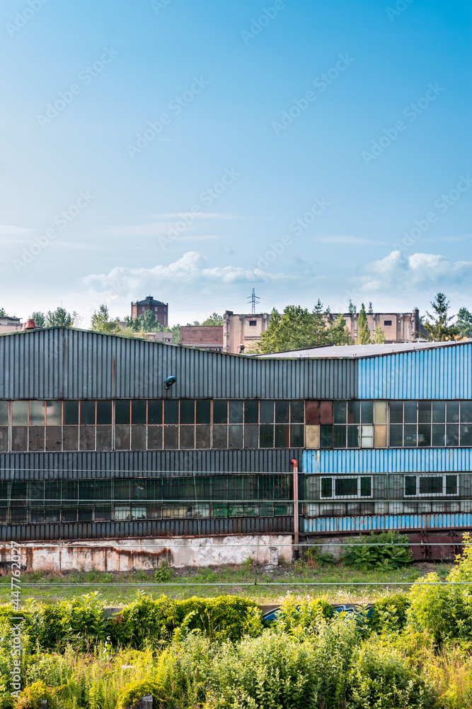 Old, metal, rusted factory hall situated in industrial district in a city. Tall grass and bushes in the foreground, cityscape and blue sky in the background. Old, metal, rusted factory hall situated in industrial district in a city. Tall grass and bushes in the foreground, cityscape and blue sky in the background.