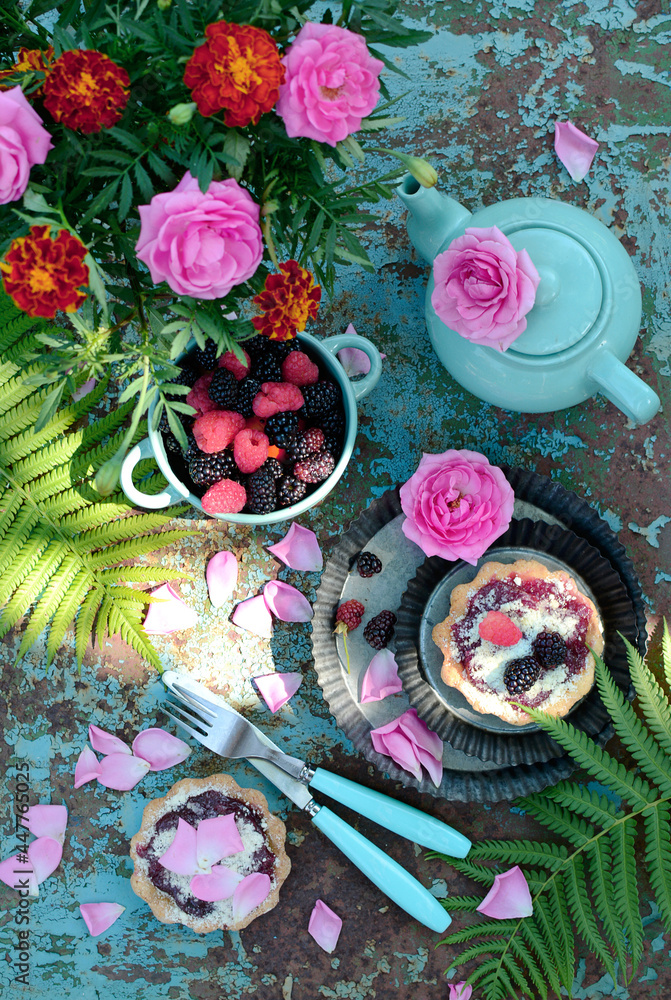 Tartlets with jam and berries, teapot, flowers and fern branches on a turquoise background. View from above