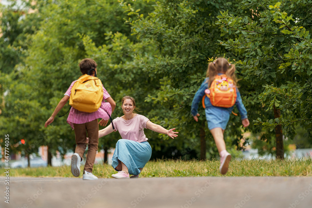Fototapeta premium loving family. caring mother meets cheerful children, boy and girl with backpacks running in her arms, meeting children hugging them, after school classes in park. Back to school. first day training