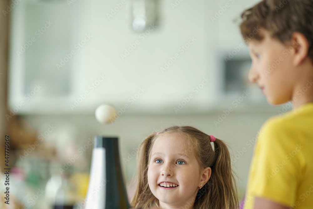 Curious boy and girl conducting a physical experiment called floating ball with a hairdryer and a plastic pink ball at home in the kitchen. Children are happy and laughing and jumping merrily