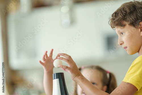 Curious boy and girl conducting a physical experiment called floating ball with a hairdryer and a plastic pink ball at home in the kitchen. Children are happy and laughing and jumping merrily