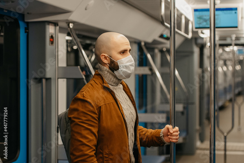 A bald man with a beard in a face mask is holding the handrail in a subway car.