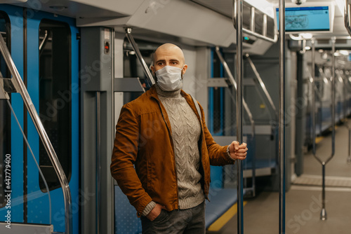 A bald man with a beard in a face mask is holding the handrail in a subway car.