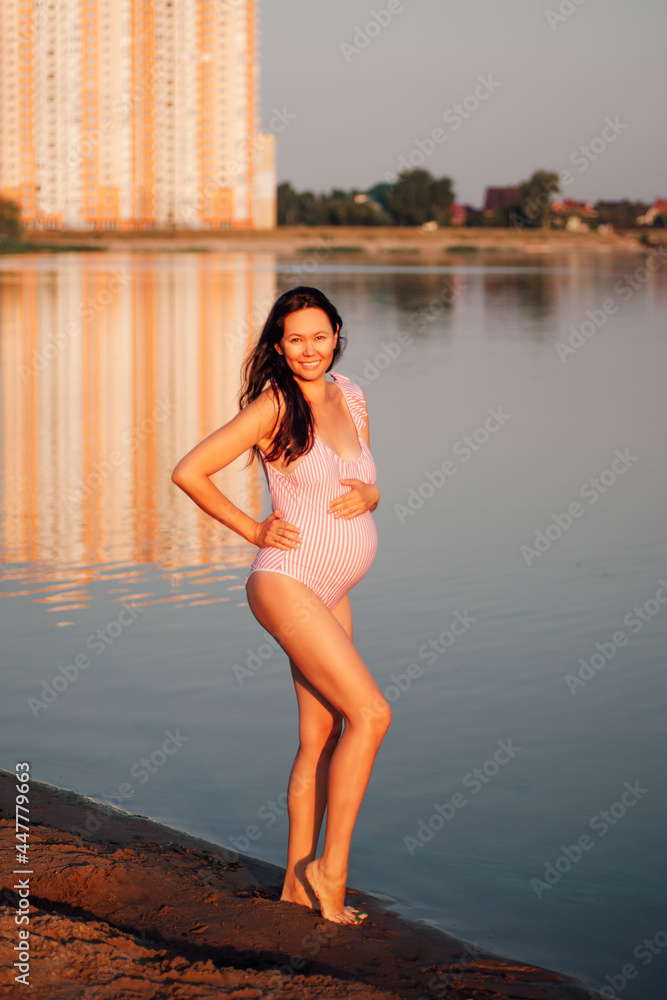 pregnant woman in a swimsuit, laughing, beautiful asian pregnant woman posing in a pink swimsuit holds her hands on her pregnant belly on a sandy beach on a summer trip. 