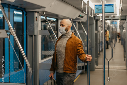 A bald man with a beard in a face mask is holding the handrail in a subway car.