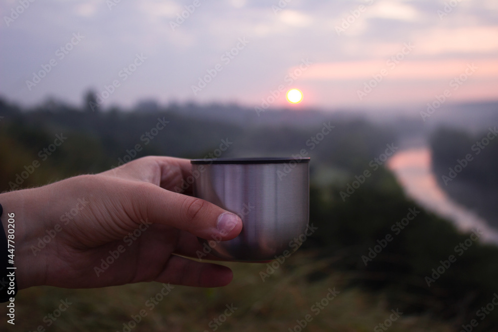 Obraz premium A man holds a metal mug in his hand in the morning against the background of dawn and the river. Travel concept