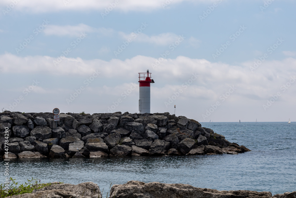 A small red and white lighthouse on a rocky pier at Lakefront Promenade Park, Mississauga, Lake Ontario. Sailboats in background. A small red and white lighthouse on a rocky pier at Lakefront Promenade Park, Mississauga, Lake Ontario. Sailboats in background.
