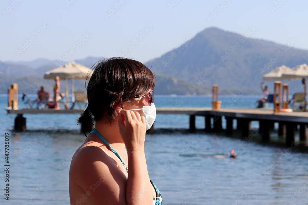 Woman in protective mask and sunglasses standing on a sea beach on pier and mountains background. Safety at tourist resort during coronavirus pandemic