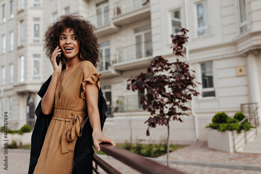 Fototapeta premium Attractive dark-skinned woman speaks on phone outside. Curly brunette lady in brown dress and black coat leans on bench outdoors.