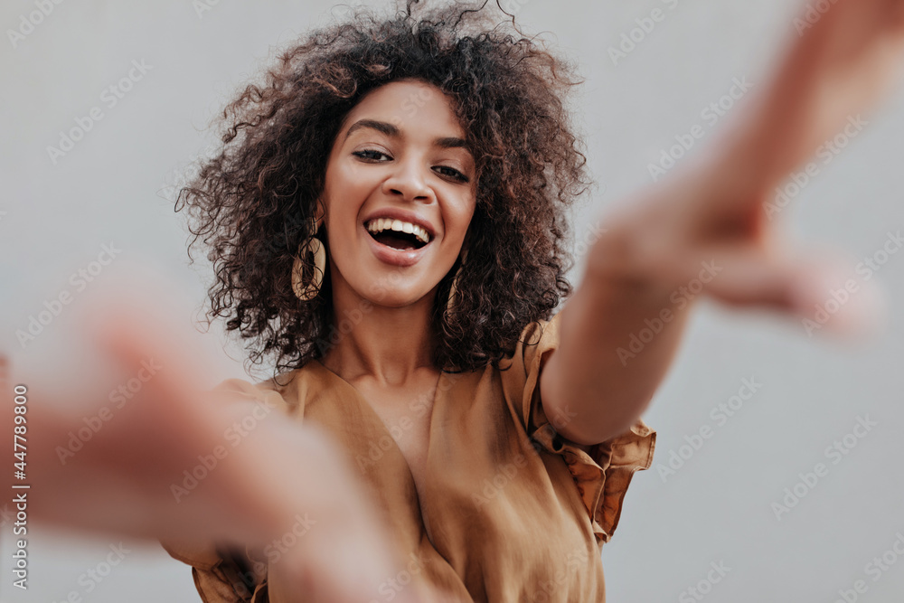 Attractive curly woman wants to touch camera. Curly dark-skinned girl in brown blouse smiles on grey background. Attractive curly woman wants to touch camera. Curly dark-skinned girl in brown blouse smiles on grey background.