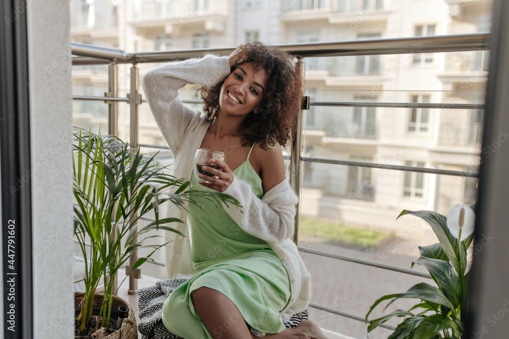 Attractive lady in silk dress holds coffee glass on balcony. Charming dark-skinned woman in white cardigan sits on terrace. Attractive lady in silk dress holds coffee glass on balcony. Charming dark-skinned woman in white cardigan sits on terrace.