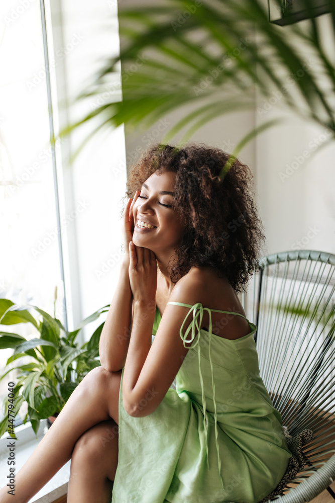 Excited brunette African woman in silk dress smiles with closed eyes in room with plants. Happy dark-skinned lady in green outfit sits on chair. Excited brunette African woman in silk dress smiles with closed eyes in room with plants. Happy dark-skinned lady in green outfit sits on chair.