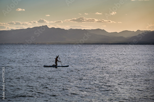 Man paddle surfing  while working on dynamic balance  on a summer sunset  on a calm sea with no waves. Improving his attitude towards everyday life and eliminating stress.