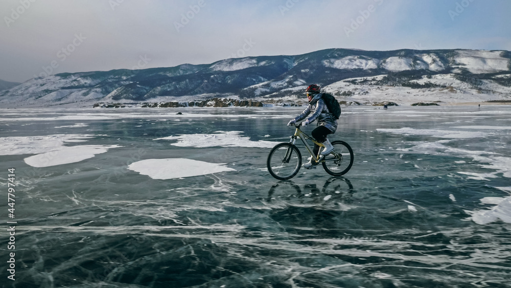 Obraz premium Woman is riding bicycle on the ice. Girl is dressed in a silvery down jacket, cycling backpack and helmet. Ice of the frozen Lake Baikal. Tires on bike are covered with spikes. Traveler is ride cycle.