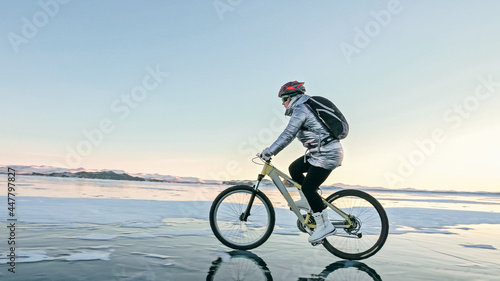 Woman is riding bicycle on the ice. Girl is dressed in a silvery down jacket  cycling backpack and helmet. Ice of the frozen Lake Baikal. Tires on bike are covered with spikes. Traveler is ride cycle.