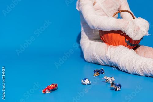 Child in mummy costume eats candies from basket in the form of pumpkin against blue background