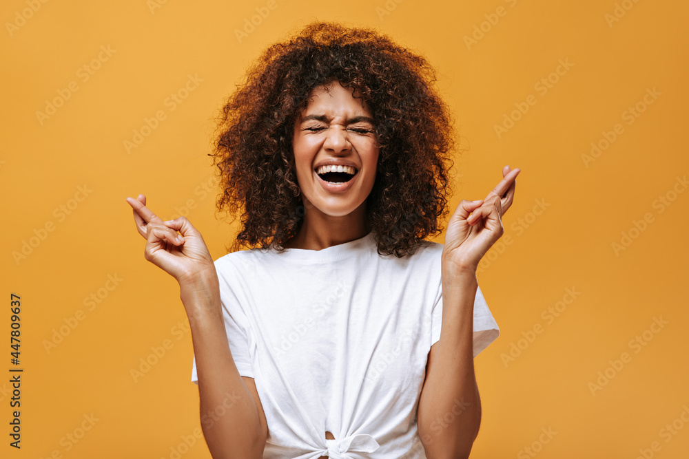 Joyful woman in light clothes crossing her fingers on yellow backdrop. Cool girl with curly hairstyle laughing on isolated background..
