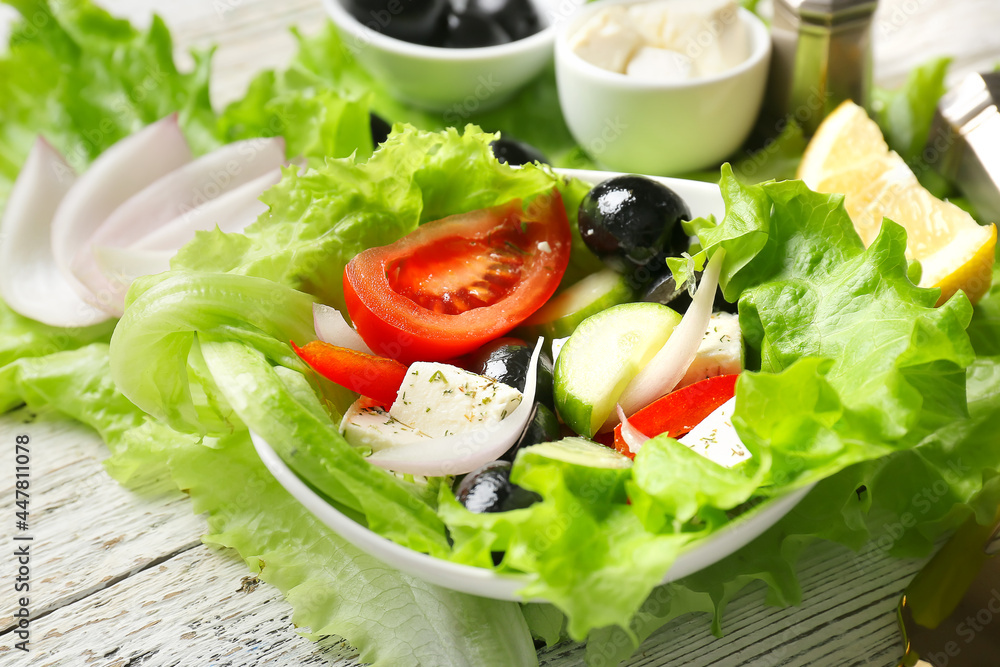 Bowl with tasty Greek salad and ingredients on light wooden background, closeup