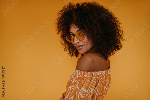Brunette woman in orange clothes looking into camera on isolated background. Cool girl with fluffy hair in sunglasses smiling on yellow backdrop..